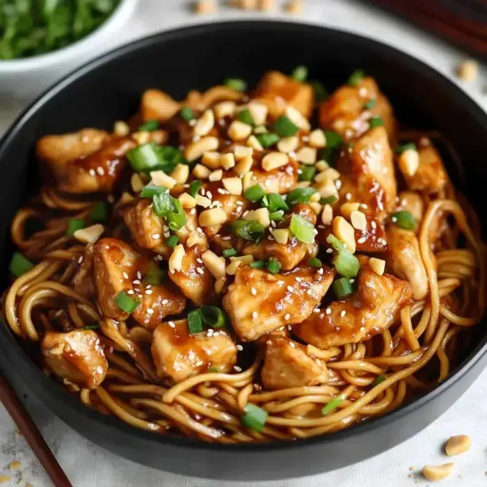 A black bowl filled with noodles, chicken pieces, green onions, crushed peanuts, and sesame seeds covered in a rich, savory sauce.