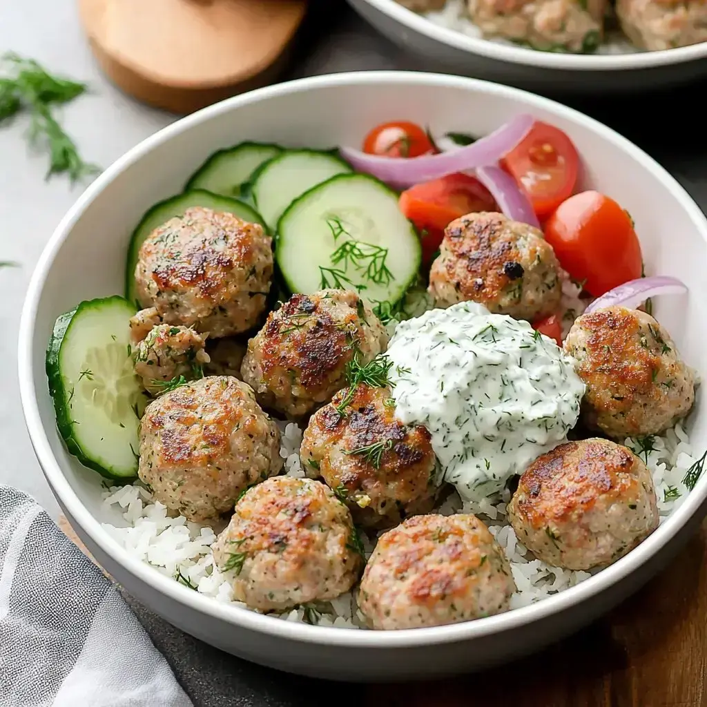 A dish of rice and juicy meatballs topped with tzatziki, cucumbers, tomatoes, red onions, and fresh herbs.