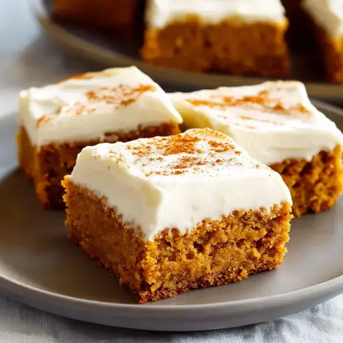 Pumpkin bars stacked on a plate with a thick layer of cream cheese frosting and a dusting of cinnamon.