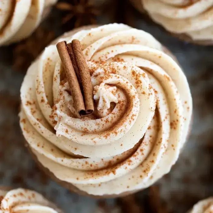 Close up of a cupcake frosted with buttercream, two cinnamon sticks, dusted cinnamon on top.