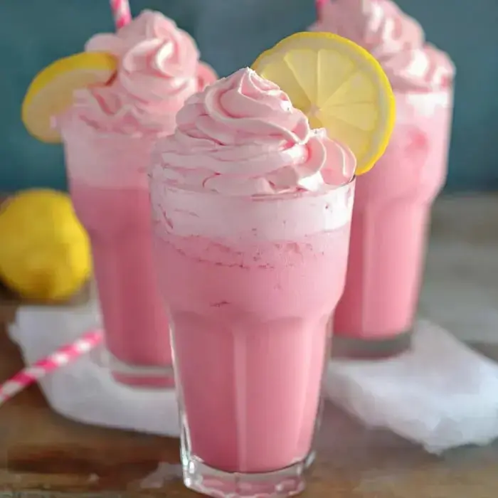 Three glasses packed with frothy pink drinks, topped with whipped cream, lemon wedges, plus a fresh lemon and straw on the side.