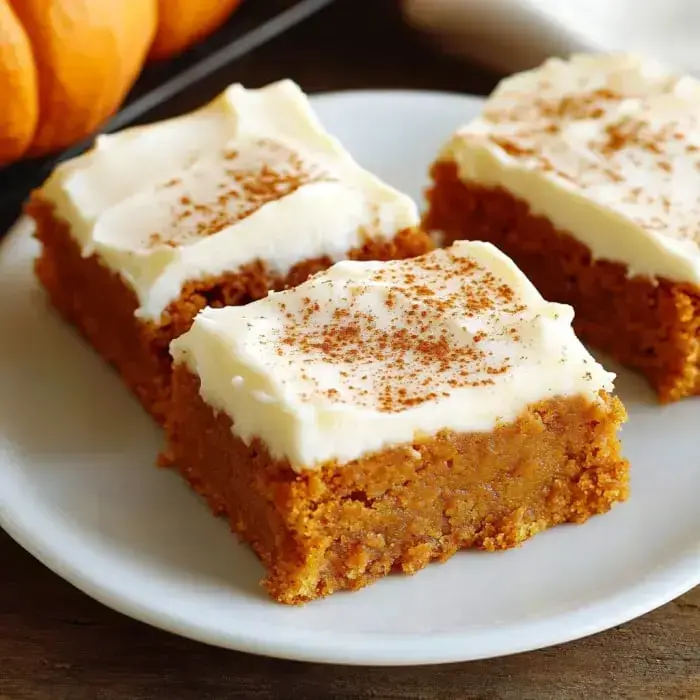 Slices of pumpkin bars with creamy frosting and cinnamon dust, set on a white plate.