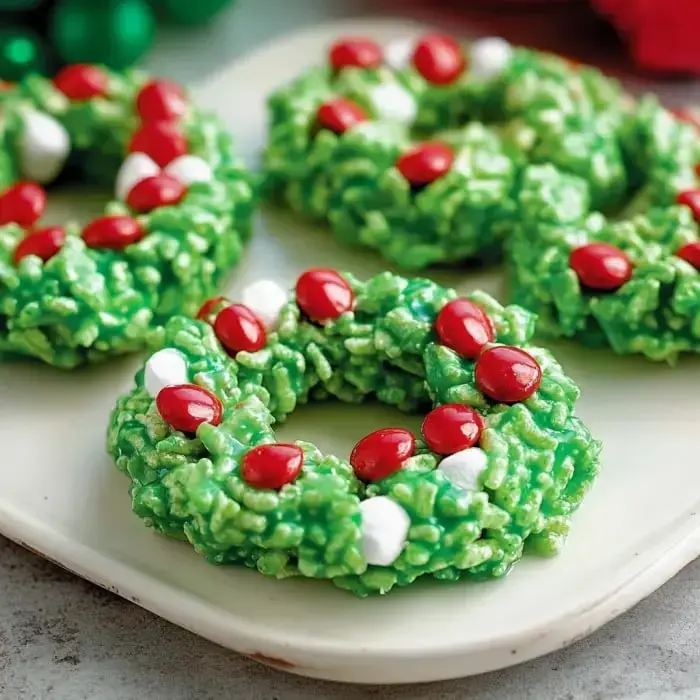 Festive green cereal treats shaped like wreaths with pops of red candy and marshmallow on a plate.