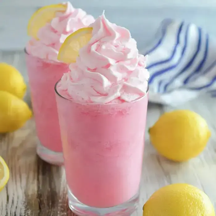 Two glasses of pink whipped drink with whipped cream and lemon wheels, next to fresh lemons on a wooden board.