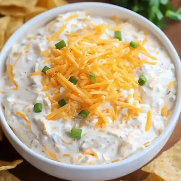 A big bowl packed with creamy dip covered in cheddar cheese and green onions, tortilla chips right alongside.