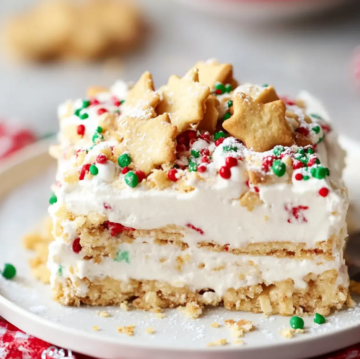 A white plate holds a slice of layered creamy dessert topped with a puff of whipped cream, sparkly holiday sprinkles, and a star cookie for a festive finish.