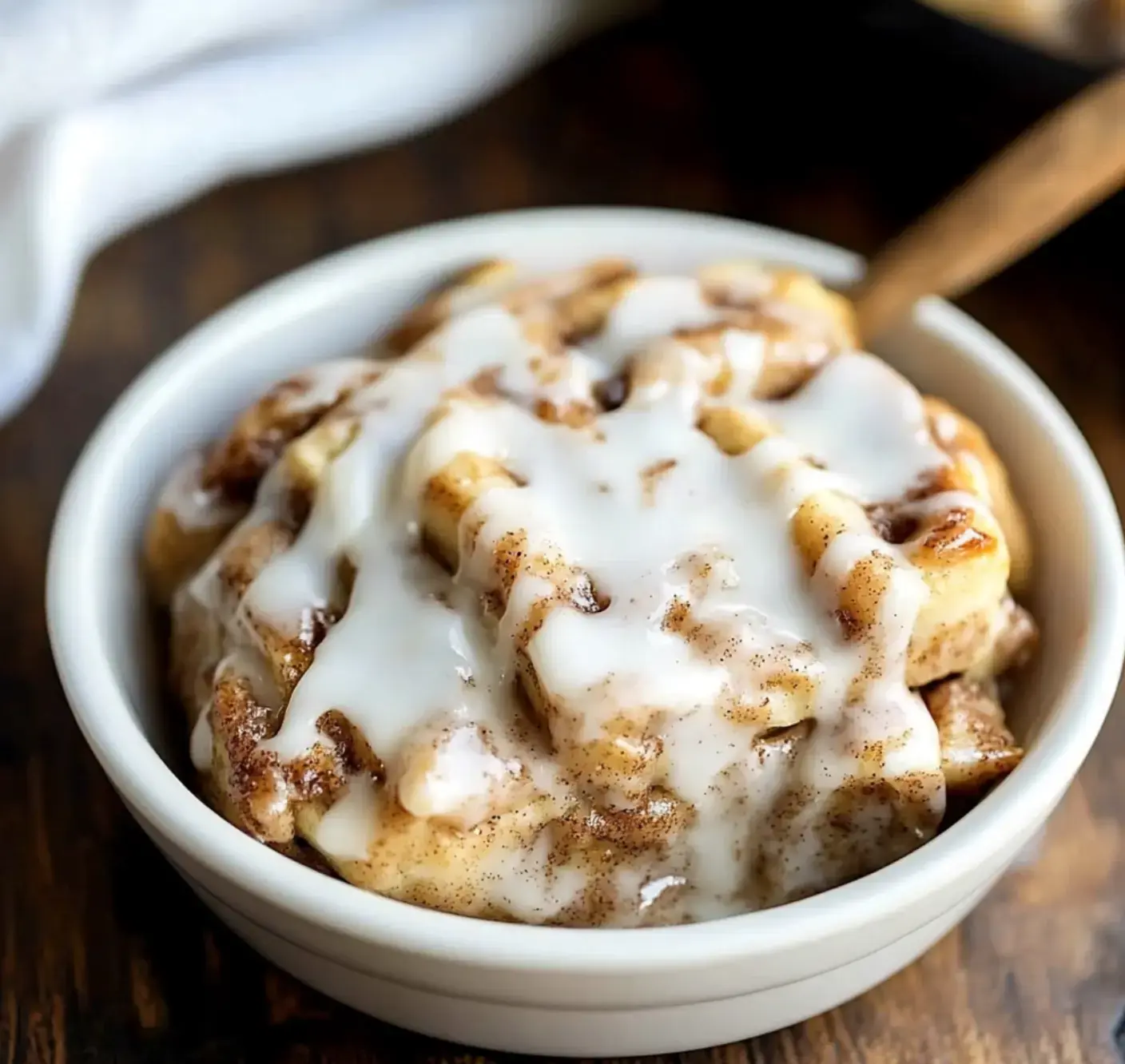 A bowl of cinnamon roll pieces topped with white icing on a wood table.