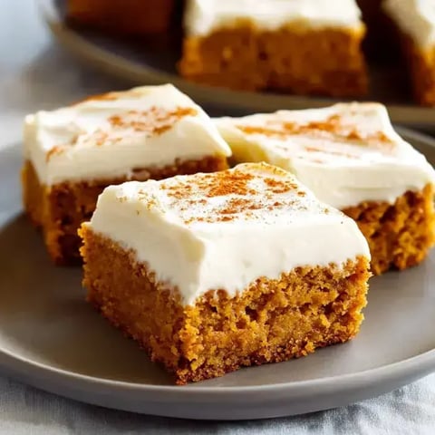 Pumpkin bars stacked on a plate with a thick layer of cream cheese frosting and a dusting of cinnamon.